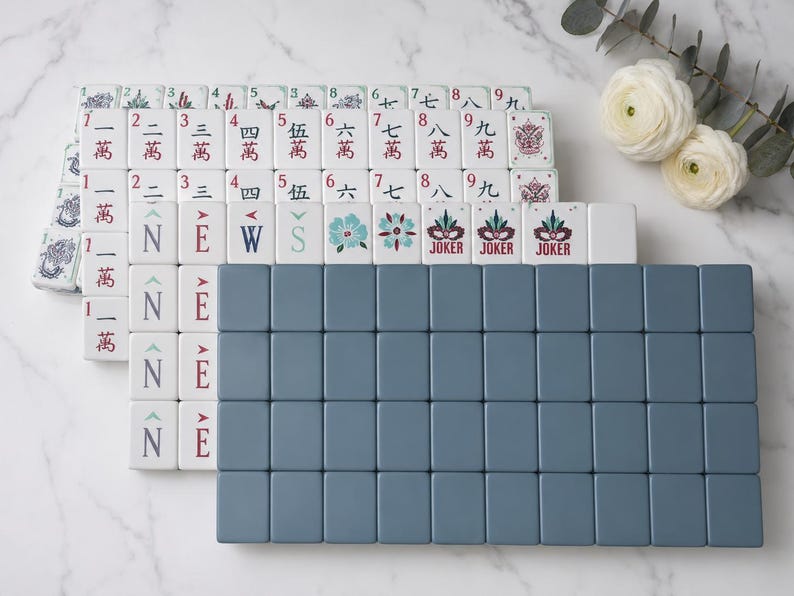 May include: A set of rectangular mahjong tiles arranged in rows. The tiles feature white faces with red, blue, and green printed Chinese characters, floral motifs, and the word JOKER. A large block of solid slate blue tiles is positioned in the front.