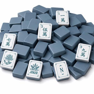 May include: A large pile of rectangular mahjong tiles with a smooth, slate-blue finish. Several tiles are face-up, displaying white surfaces with green Chinese characters, a floral illustration, the word JOKER, and the letter N with arrows.