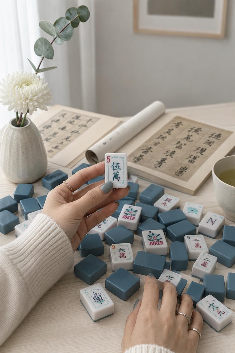 May include: A set of Mahjong tiles featuring white faces with black and red Chinese characters, floral designs, and the word JOKER, alongside solid blue-grey tiles. One white tile with the number 5 and Chinese characters is held up.