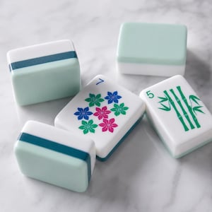 Puede incluir: Cinco fichas de mahjong rectangulares con superficies lisas. Dos fichas son de color verde claro liso con una franja verde azulado oscuro. Una ficha presenta un grupo de flores azules, rosas y verdes con el número 7. Una ficha muestra tallos de bambú verdes y el número 5.