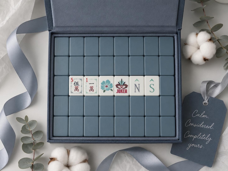 May include: A set of blue mahjong tiles arranged in a rectangular box. Six tiles in the center feature unique designs including Chinese characters, a blue flower, a red joker mask, and the letters N and S. A blue tag reads Calm. Considered. Completely yours.