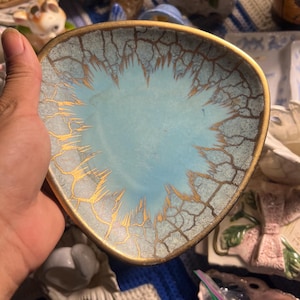 Puede incluir: Un plato triangular de cerámica con un centro azul claro que se transforma en un patrón texturizado y agrietado de azul claro y blanco. Los bordes del plato tienen un acabado con un borde dorado metálico, resaltando el diseño irregular y dentado que irradia desde el centro hacia el borde dorado.