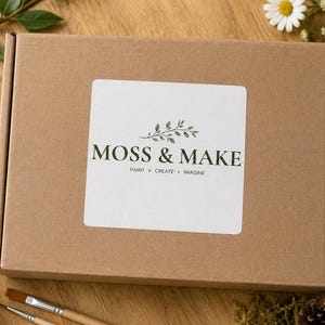Może przedstawiać: Prostokątne brązowe pudełko kartonowe z białą kwadratową etykietą zawierającą napis MOSS & MAKE oraz hasło PAINT &bull; CREATE &bull; IMAGINE, wraz z małą ilustracją zielonych liści wyśrodkowaną nad nazwą marki.