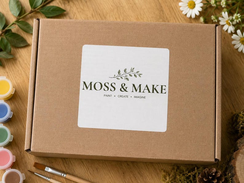 Może przedstawiać: Prostokątne brązowe pudełko kartonowe z białą kwadratową etykietą zawierającą napis MOSS & MAKE oraz hasło PAINT &bull; CREATE &bull; IMAGINE, wraz z małą ilustracją zielonych liści wyśrodkowaną nad nazwą marki.