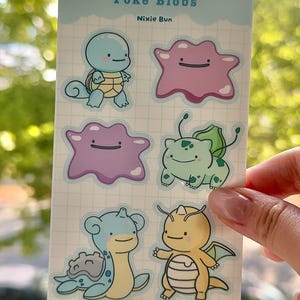 May include: A rectangular sticker sheet titled Poké Blobs by Nixie Bun featuring six cartoon character illustrations. The designs include a light blue turtle, a pink blob, a purple blob, a light green creature, a blue creature, and an orange dragon.
