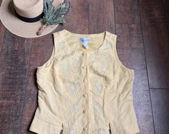 May include: A sleeveless, pale yellow vest with floral embroidery and button closures. A straw hat with a black band and a sprig of lavender are also present. The vest has a label that reads "Dara South".