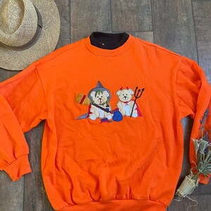 May include: Orange sweatshirt with a black collar and an embroidered design of two teddy bears dressed as a witch and a devil for Halloween.