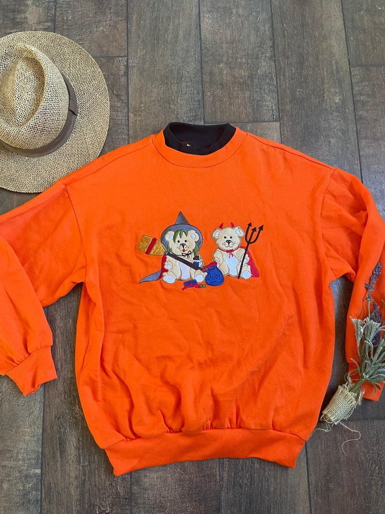 May include: Orange sweatshirt with a black collar and an embroidered design of two teddy bears dressed as a witch and a devil for Halloween.