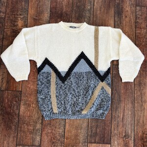 May include: A white knitted sweater with a geometric pattern in black, gray, and brown. The sweater has a crew neck and long sleeves.