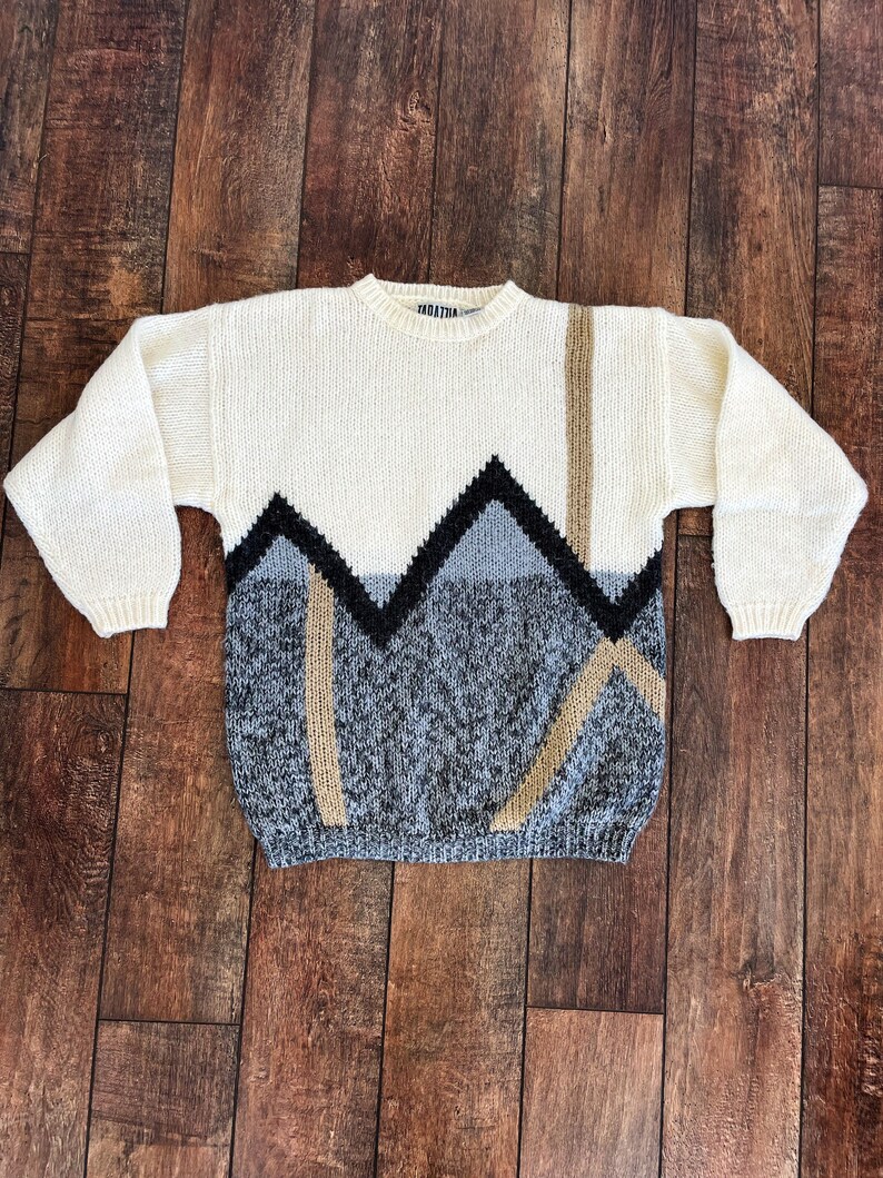May include: A white knitted sweater with a geometric pattern in black, gray, and brown. The sweater has a crew neck and long sleeves.