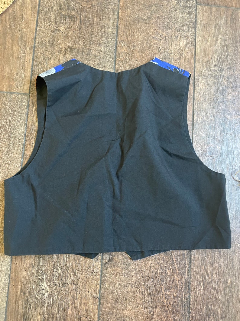 May include: Black vest with a contrasting blue and white patterned fabric on the shoulders. The vest is made of a solid black material and has a classic, tailored design. The back of the vest is visible, showing the full shape and construction.