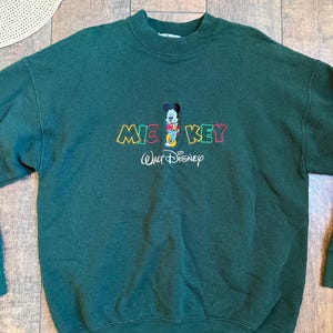 May include: Dark green sweatshirt featuring a Mickey Mouse graphic. The design includes the word "MICKEY" in colorful letters and the "Walt Disney" logo. A straw hat is visible in the background.