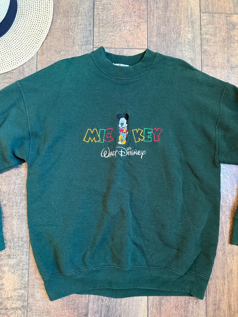 May include: Dark green sweatshirt featuring a Mickey Mouse graphic. The design includes the word "MICKEY" in colorful letters and the "Walt Disney" logo. A straw hat is visible in the background.