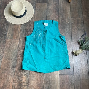 以下が含まれることがあります： ボタン留めのフロントとシルバーとホワイトの刺繍デザインが施された、ターコイズブルーのノースリーブトップ。トップは軽量な生地で作られており、ゆったりとしたフィット感です。
