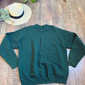 May include: A dark green sweatshirt with long sleeves. A straw hat with a black band and a sprig of lavender are next to the sweatshirt. The items are on a wooden surface.