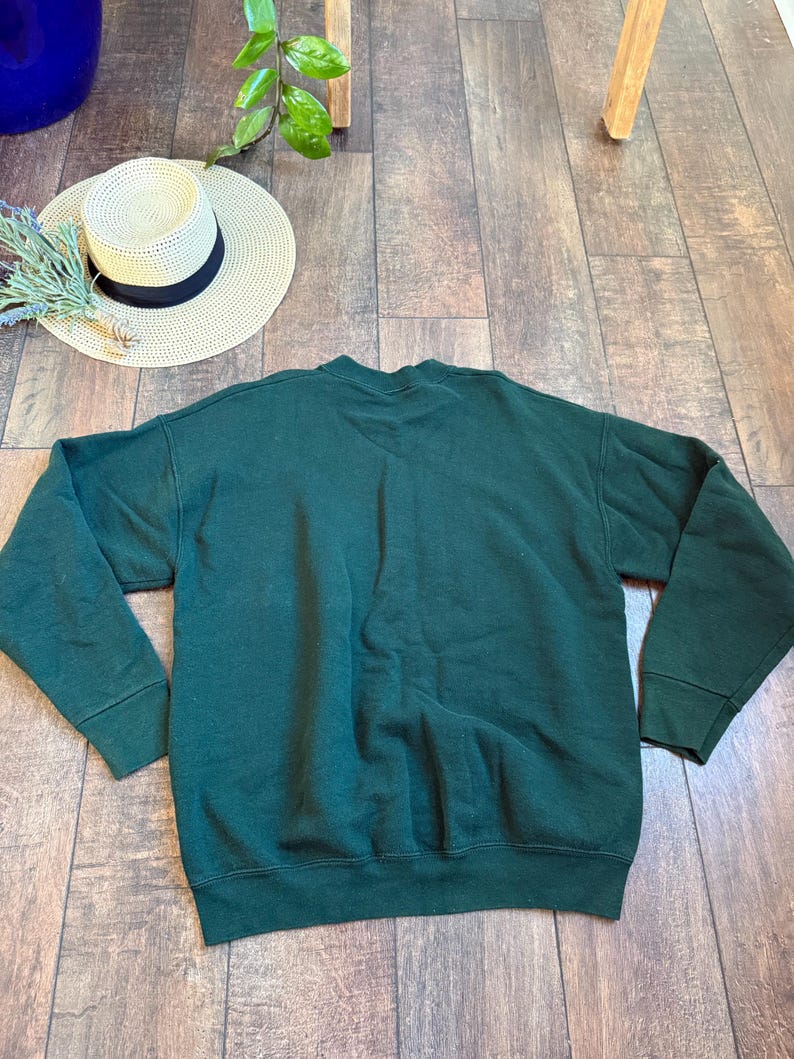 May include: A dark green sweatshirt with long sleeves. A straw hat with a black band and a sprig of lavender are next to the sweatshirt. The items are on a wooden surface.