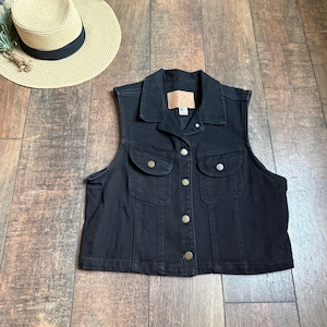 Könnte beinhalten: Eine schwarze Jeansweste mit Kragen, zwei Brusttaschen und Knopfverschlüssen. Ein Strohhut mit schwarzem Band liegt in der Nähe. Die Weste hat einen Vintage-Look.