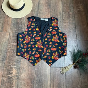 May include: A black sleeveless vest with a fall-themed print featuring pumpkins, leaves, and sunflowers. The vest has a button closure and a pointed hem.