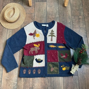 Op de afbeelding: Een blauwe cardigan met een patchwork ontwerp met verschillende bosdieren en planten. De cardigan heeft een knoopsluiting en lange mouwen.