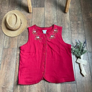 以下が含まれることがあります： 刺繍された鳥と花のデザインが施された、赤いノースリーブのボタン留めベスト。ベストは、波状の裾とボタン留めになっています。