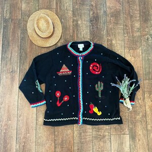 May include: Black cardigan sweater with colorful embroidered details, including a sombrero, maracas, a cactus, and a chili pepper. The sweater has a blue and white trim and a button closure.