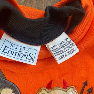 May include: Orange sweatshirt with a black collar and a small tag that reads "basic Editions". The sweatshirt has an embroidered design of a teddy bear with devil horns and a pitchfork.