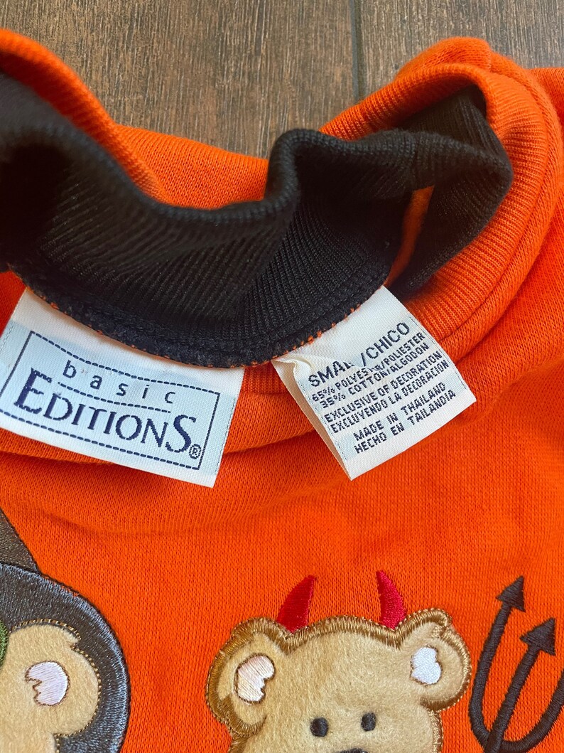 May include: Orange sweatshirt with a black collar and a small tag that reads "basic Editions". The sweatshirt has an embroidered design of a teddy bear with devil horns and a pitchfork.