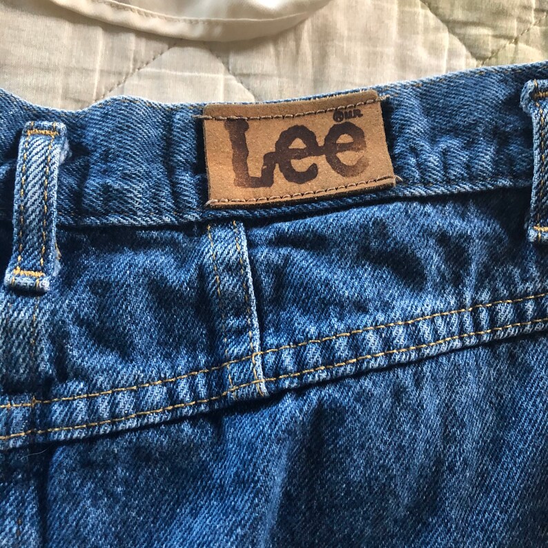 May include: A close-up of a pair of blue denim jeans with a brown leather patch that says "Lee" on the back waistband.