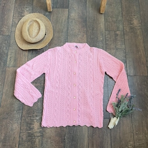 May include: A pink crocheted cardigan with a button-up front and a scalloped hem. The cardigan has a cable knit pattern and pearl-like buttons.
