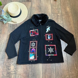 Puede incluir: Cárdigan negro con cremallera, cuello de piel sintética y parches con temática invernal. Los parches presentan imágenes de patines de hielo, muñecos de nieve y copos de nieve, enmarcados con bordes de cuadros rojos y verdes. También se ve un sombrero de paja con una banda negra.