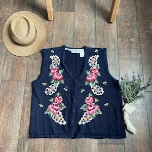 May include: A navy blue sleeveless knitted vest with a floral pattern in shades of pink, purple, white, and green. The vest has a V-neck and button closure.