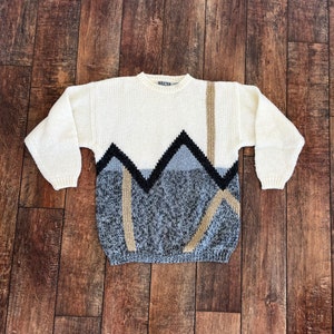 May include: A white knitted sweater with a geometric pattern in black, gray, and beige. The pattern features a series of triangles and lines.