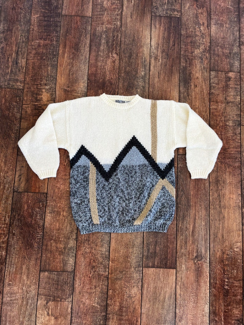 May include: A white knitted sweater with a geometric pattern in black, gray, and beige. The pattern features a series of triangles and lines.