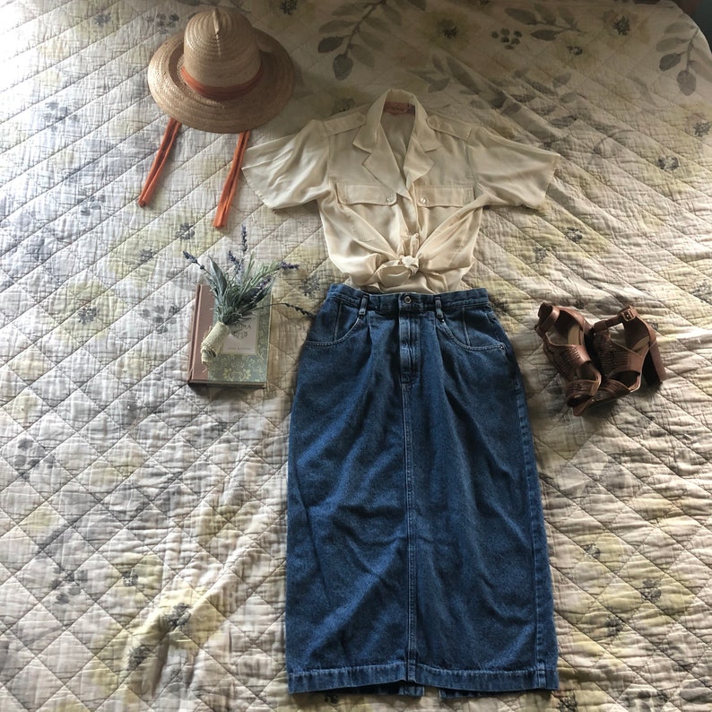 May include: A light blue button-up shirt with a white background and a denim skirt. The skirt has a high waist and a straight cut. The outfit is laid out on a white quilted bedspread. There are two brown sandals on the bedspread.