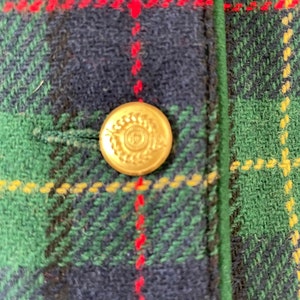 May include: A close-up of a gold button with a laurel wreath design on a green, red, and blue plaid fabric.