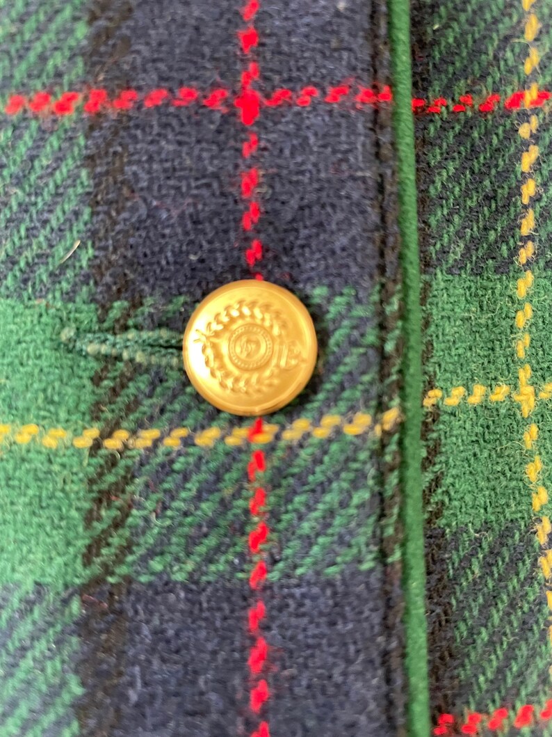 May include: A close-up of a gold button with a laurel wreath design on a green, red, and blue plaid fabric.