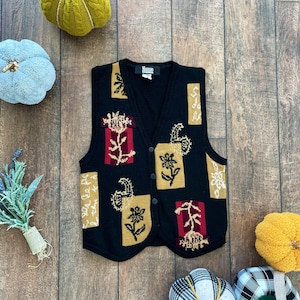 May include: Black knitted vest with a floral pattern in shades of gold, red, and white. The vest has a V-neck and button closure. The brand name "Mossimo" is visible on the label.