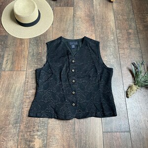 以下が含まれることがあります： 花柄とゴールドのボタンが付いた、黒いノースリーブベスト。ベストはVネックで、柔らかく質感のある生地で作られています。ベストには「Laura Scott」のラベルが付いています。