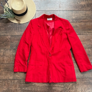 May include: A bright red blazer with a single-button closure and two front pockets. A straw hat with a black band and a bundle of lavender are next to the blazer. The blazer is a solid color and has a classic design.