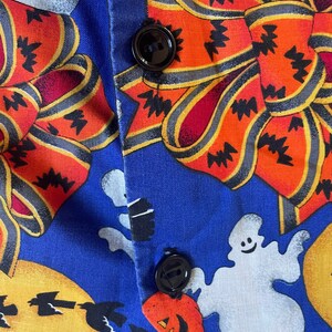 May include: Close-up of a blue Halloween-themed fabric with orange bows, ghosts, jack-o'-lanterns, and bats. The fabric is buttoned with two black buttons. The bows have black zig-zag patterns.