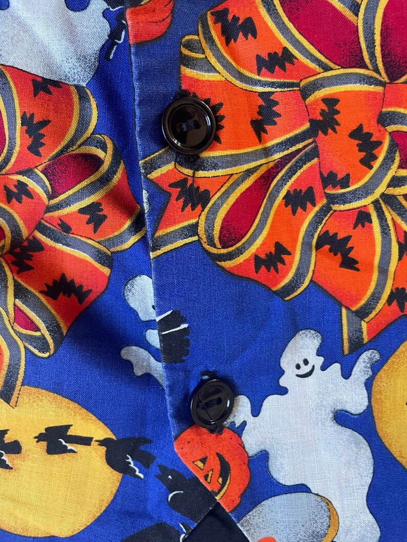 May include: Close-up of a blue Halloween-themed fabric with orange bows, ghosts, jack-o'-lanterns, and bats. The fabric is buttoned with two black buttons. The bows have black zig-zag patterns.