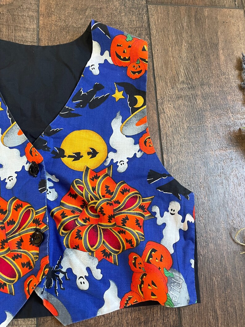 May include: A blue Halloween vest with a black back and buttons. The vest features a festive print of orange jack-o'-lanterns, white ghosts, bats, and a large orange bow. The vest is a fun and unique addition to any Halloween costume.