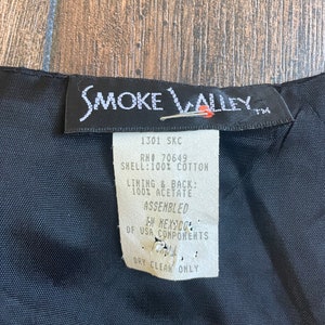 Może przedstawiać: Czarna metka odzieżowa marki Smoke Valley z białym tekstem. Metka zawiera tekst "1301 SKC", "RN# 70649", "SHELL: 100% COTTON", "LINING & BACK: 100% ACETATE", "ASSEMBLED", "IN MEXICO", "OF USA COMPONENTS" i "DRY CLEAN ONLY".