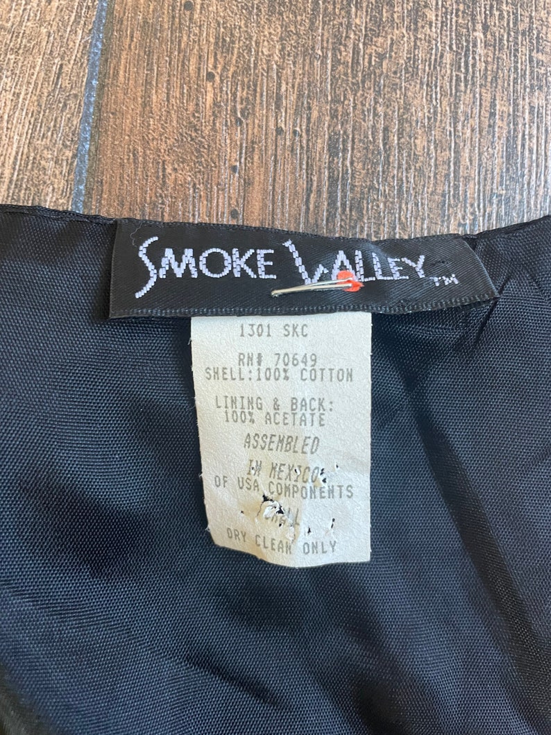 Może przedstawiać: Czarna metka odzieżowa marki Smoke Valley z białym tekstem. Metka zawiera tekst "1301 SKC", "RN# 70649", "SHELL: 100% COTTON", "LINING & BACK: 100% ACETATE", "ASSEMBLED", "IN MEXICO", "OF USA COMPONENTS" i "DRY CLEAN ONLY".