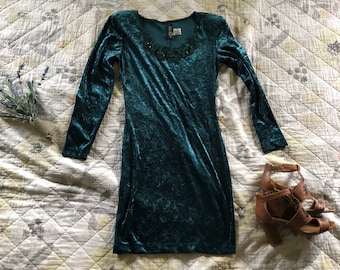 May include: A teal blue velvet dress with long sleeves and a beaded neckline. The dress is photographed on a white quilted fabric.