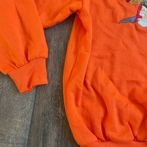 May include: Orange sweatshirt with a cartoon character embroidered on the front. The character is holding a bottle and a sword.