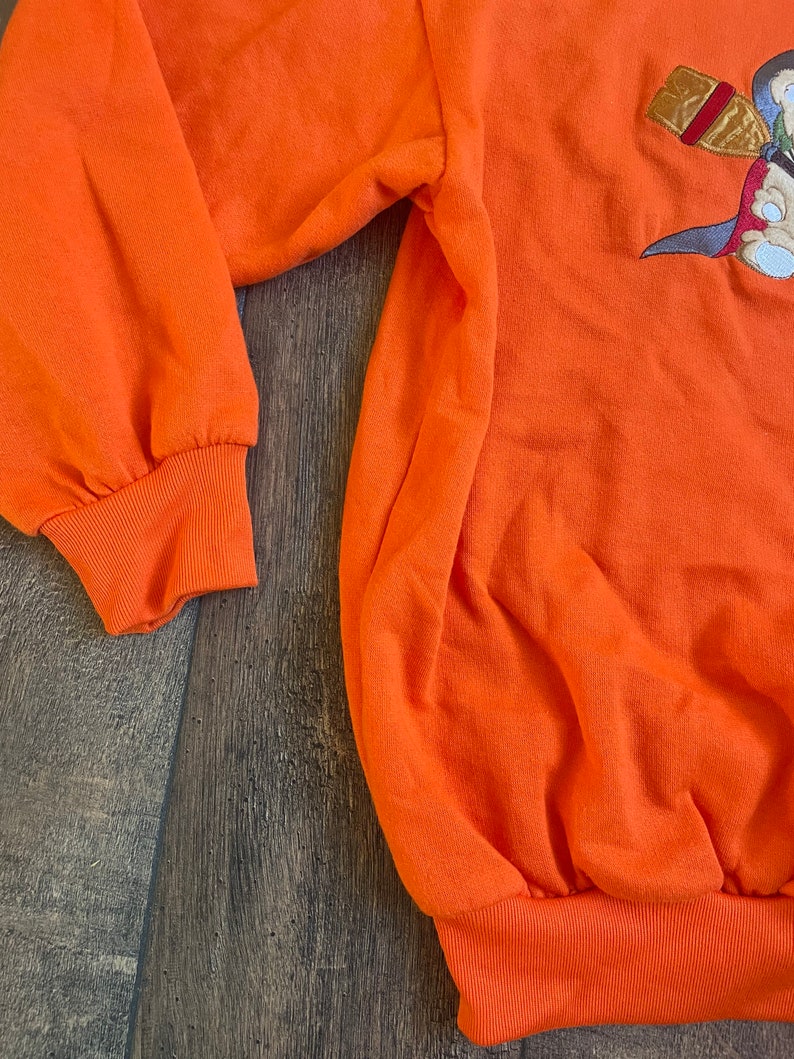 May include: Orange sweatshirt with a cartoon character embroidered on the front. The character is holding a bottle and a sword.
