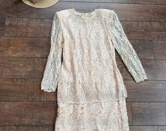 Könnte beinhalten: Ein zweiteiliges Vintage-Spitzenkleid in hellem Pfirsichton. Das langärmlige Oberteil und der Rock sind mit einem floralen Spitzendesign bedeckt. Ein Strohhut mit schwarzem Band und ein Zweig Lavendel sind ebenfalls im Bild.