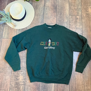 May include: A dark green sweatshirt featuring the word "MICKEY" in colorful embroidery, with a cartoon Mickey Mouse graphic. The Walt Disney logo is embroidered below. A straw hat with a black band and a sprig of dried flowers are also visible.