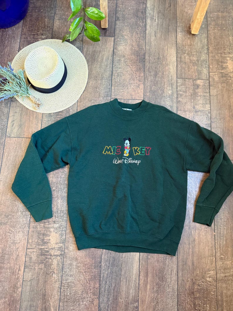 May include: A dark green sweatshirt featuring the word "MICKEY" in colorful embroidery, with a cartoon Mickey Mouse graphic. The Walt Disney logo is embroidered below. A straw hat with a black band and a sprig of dried flowers are also visible.
